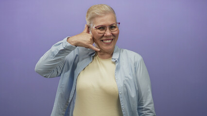 Fototapeta premium Woman pointing finger and making call me hand gesture in purple studio wearing glasses and casual shirt; confidence.
