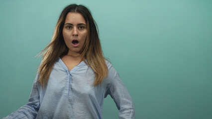 Obraz premium Woman mouth open hand raised in mint green studio showing open mouth and expressive gesture, wearing light blue blouse with loose sleeves; surprise reaction.