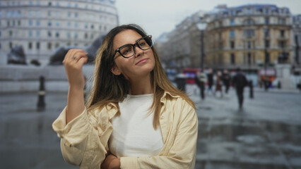 Obraz premium Woman wearing glasses touches her hair and looks up on a rainy city street near historic buildings, casual shirt and ring visible; contemplation.