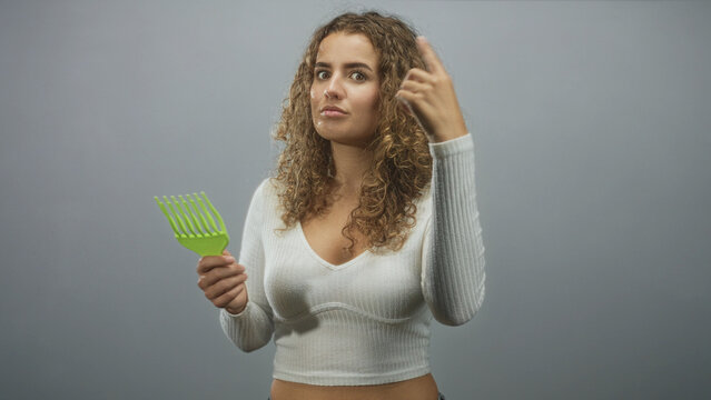 Woman with curly hair points finger while holding a green wide tooth comb and wearing a white crop top in a studio setting; confidence self care.