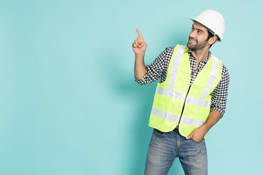 A smiling male engineer in a white hard hat and safety vest points his finger up and looks at copy space, isolated on a cyan background. Great for industrial construction and advertising concepts