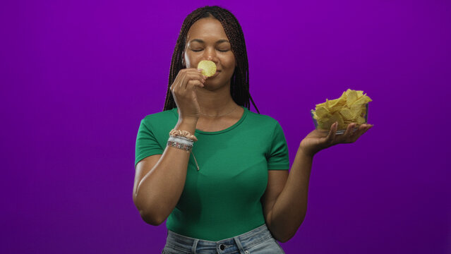 Woman holding a glass bowl of potato chips, hand reaching to grab a chip in studio with purple backdrop and green shirt; snacking pleasure everyday.