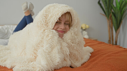 Woman lying on bed wrapped in a fluffy cream blanket, smiling with face and bare cheek visible in a bright studio  cozy contentment. © Krakenimages.com