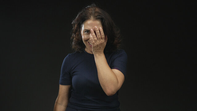Middle age woman covers face with hand, smiling and laughing in studio with dark backdrop and navy tshirt; playful joy.