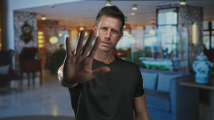 Fototapeta premium Man in black t shirt with hand palm forward to camera stop in hotel building lobby; defiance boundary privacy.