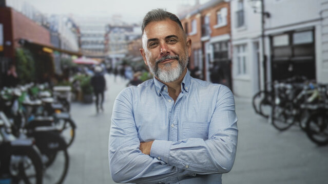 Middle aged man with arms crossed smiling on a busy street lined with bicycles and brick buildings; approachable confidence.