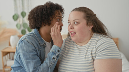 Obraz premium Woman listens as man whispers into her ear, hand near her face on a bedroom bed with plants and nightstand visible; secret affection playful.