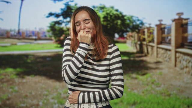 Woman biting fingernail with hand to mouth, eyes closed, striped top visible, standing near stone fence and park lawn on street; nervousness vulnerability.