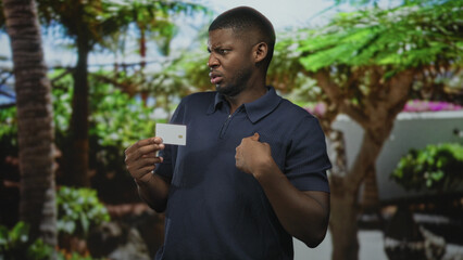 Fototapeta premium Man holds white creditcard with visible chip and points finger to chest in a building courtyard with palm trees and plants; doubt identity.