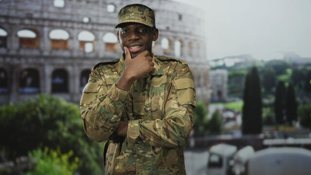 Man in camouflage military uniform and cap touches chin with hand while standing before a historic building; pride duty.