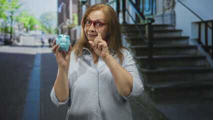 Fototapeta premium Senior hispanic woman holds blue piggy bank and points finger toward viewer on a street beside building steps; savings caution.