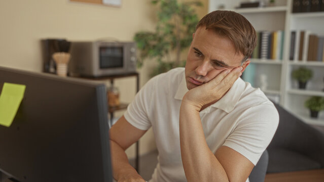 Man with hand on cheek staring at computer monitor in office building, slumped at desk with idle keyboard and pensive posture; boredom slump.