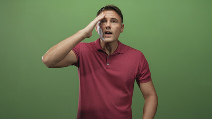 Fototapeta premium Man touching forehead with hand in studio green backdrop, wearing maroon polo and standing midshot; frustration.