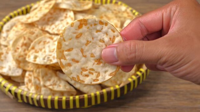 A hand picking up a piece of round tempeh chips from a bamboo woven tray on a wooden surface