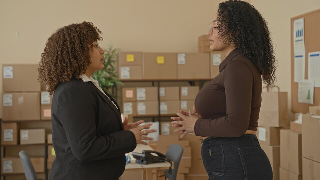 Two women small business workers clasp hands and stand with arms crossed amid stacked shipping boxes and parcels in a storage room inside a building; confidence teamwork.