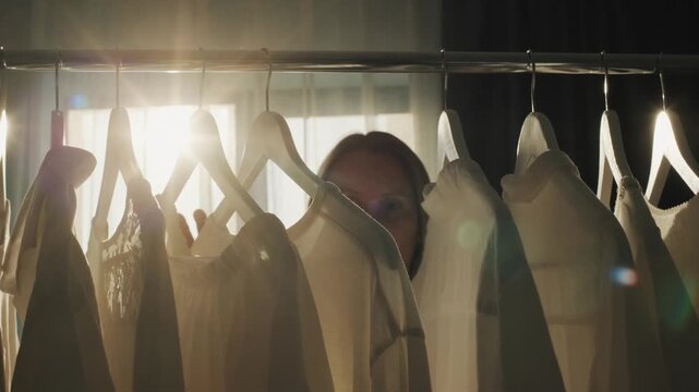 Woman choosing white clothes from a wardrobe rack in soft sunlight.