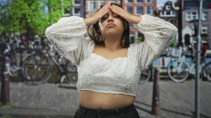 Fototapeta premium Woman in white lace top showing midriff while bringing hands to forehead on a city street with bicycles and canal; frustration reflection solitude.