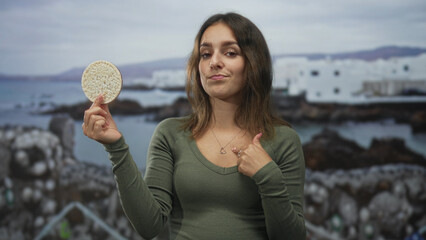 Naklejka premium Woman holding a rice cake in left hand, finger pointing to heart necklace on seaside street; confidence.