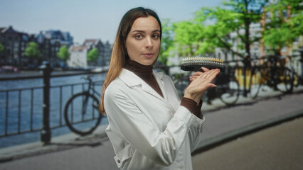 Fototapeta premium Woman dentist in white lab coat points to a dental shade guide on street by amsterdam canal, demonstrating shade selection; dental care concentration.