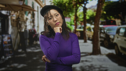 Fototapeta premium Young asian woman wearing beret and glasses, hand on chin in a purple sweater on a city street; thoughtful reflection.