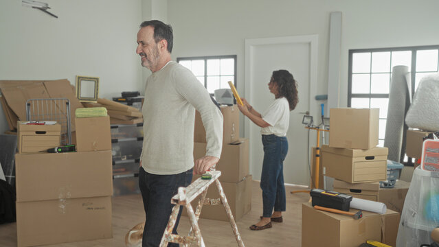 Man and woman pack stacked cardboard boxes and seal tape while man holds a ladder and woman uses a tape dispenser, moving supplies and dog in building; hopeful new beginning.