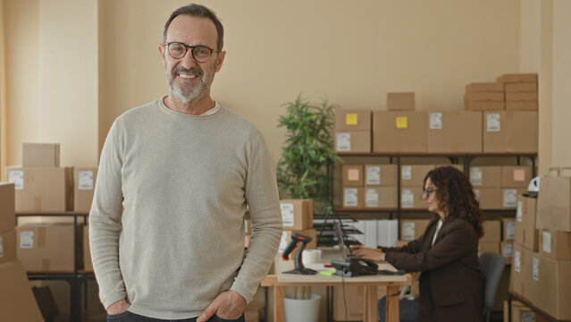 Middle-aged man smiling with hands in pockets beside stacked parcels, barcode scanner and laptop in a small business building; confidence growth.