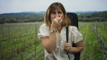 Obraz premium Young woman grimacing with hand to nose and holding a bag strap in a forest vineyard scene; disgust.