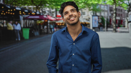 Obraz premium Man in blue shirt wearing glasses tilts his head, smiles and looks up amid cafe lights and trees on a street lined with buildings; optimism curiosity.