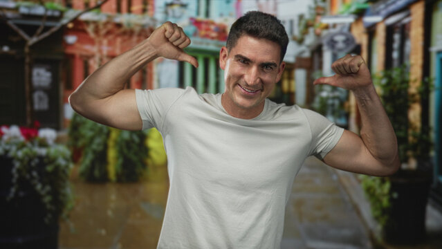 Man pointing thumbs at his chest on a narrow city street, smiling and flexing biceps in a white t shirt; confidence attitude.