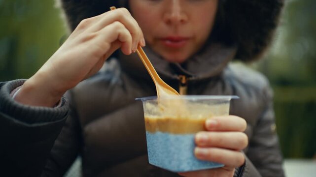 A woman wearing a dark jacket holds a small plastic cup filled with chia seed pudding while using a wooden spoon to scoop from the top