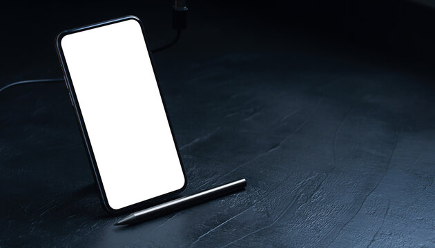 Smartphone mockup stands on a dark textured desk with a stylus pen and charging cable, featuring a blank white screen and copy space for mobile app design and digital presentation