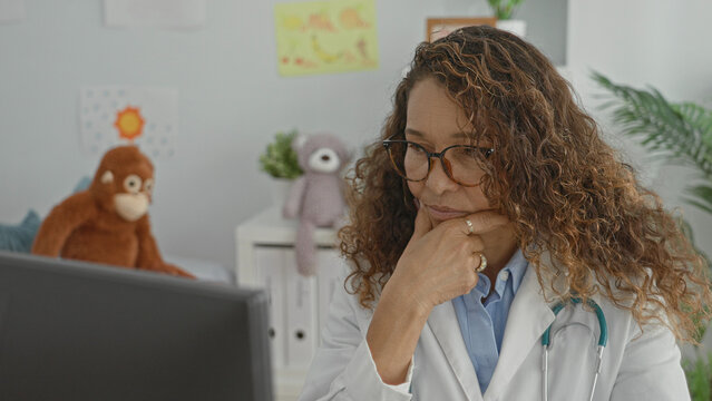 Woman doctor wearing glasses and white coat with stethoscope at computer, hand on chin, surrounded by stuffed toys and children's drawings in clinic building; focus.