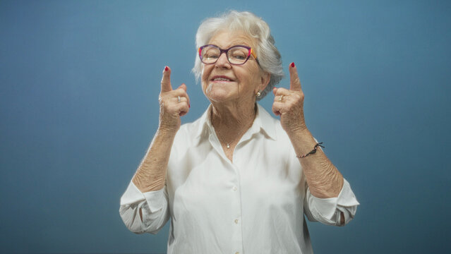 Senior woman gestures with index fingers to her cheeks then points both index fingers upward, wearing colorful glasses and white blouse in studio; playful joy.