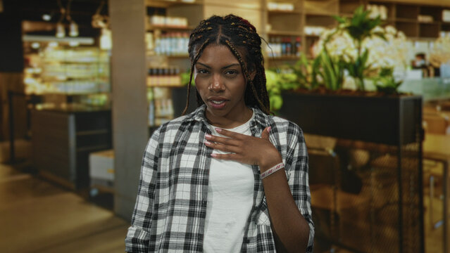 African american woman in flannel shirt with hand on chest, open mouth and braided hair standing in a building restaurant interior near plants and shelves; surprise sincerity apology.