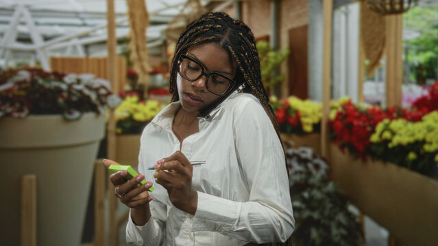 Woman with phone tucked between shoulder and ear tapping a green notepad in a building flower shop; concentration multitasking.