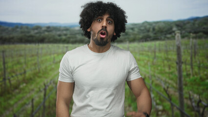 Obraz premium Man with hand to nose and arm on hip wearing white t shirt in forest vineyard field outdoors facing camera with a curious expression; curiosity reaction moment surprise.