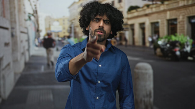Man points finger directly at camera on busy city street near bollard and parked motorcycles;accusation anger confrontation.