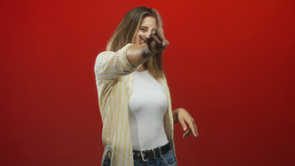 Fototapeta premium Woman showing rock hand gesture with tongue out, white tank top and jeans in a studio red backdrop; playful rebellion.