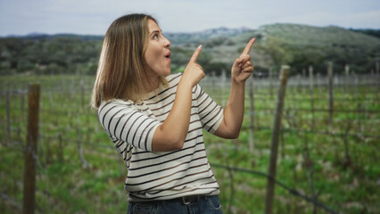 Obraz premium Woman pointing fingers to the right in a vineyard among trellis posts, smiling with visible hands and casual striped top; joy.
