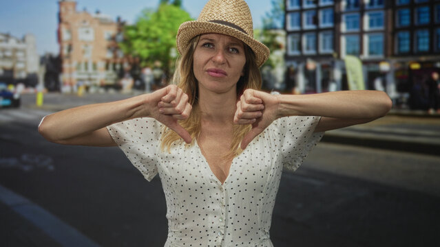 Woman showing a thumbs down gesture on a city street wearing a straw hat and polka dot dress with visible cleavage and neutral expression; disapproval.