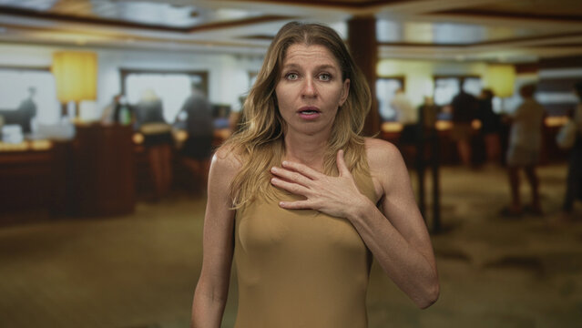 Woman clutch chest in building lobby reception area, hand pressed to chest and mouth open in a visible gasp and startled look; shock concern.