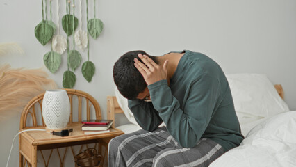 Obraz premium Man sitting on bed holding head with hands and rubbing temples beside bedside lamp in a bedroom building; stress coping.