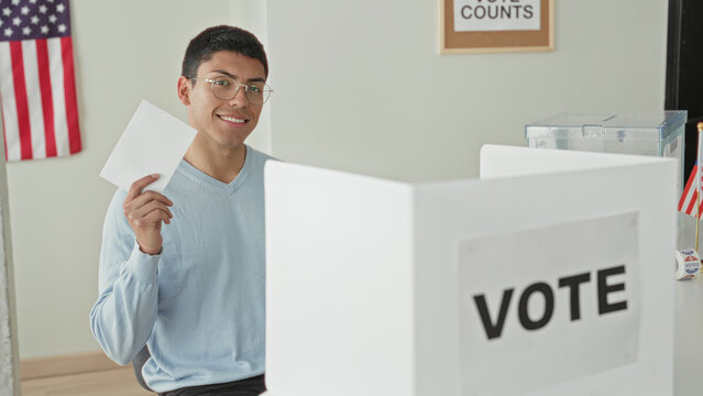 Man holding ballot in hand at voting booth inside building, smiling and raising paper; civic duty pride.