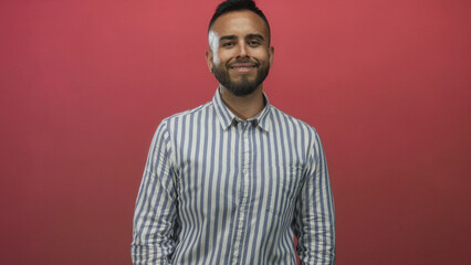 Obraz premium Man waving right hand in studio with red backdrop and blue white striped shirt, smiling at camera; friendly greeting.