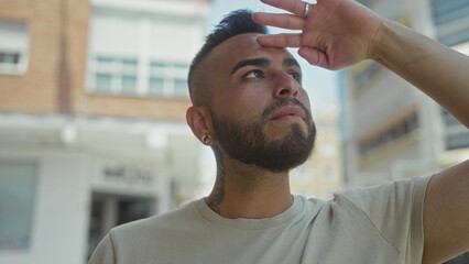 Fototapeta premium Man shielding eyes with hand on a sunny street while looking upward toward the buildings and sky; thoughtful reflection.