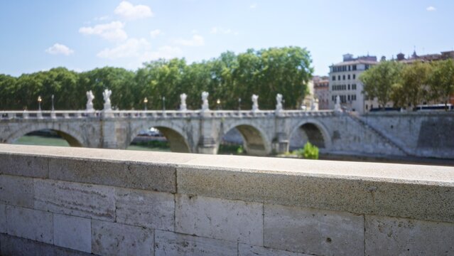 Stone parapet in sharp focus with historic roman bridge and riverside trees softly blurred in shallow bokeh street; background backplate copyspace calm.