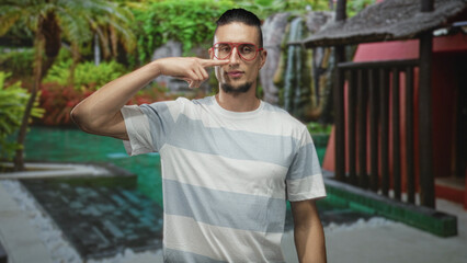 Fototapeta premium Man in striped tshirt points finger to nose and wears red glasses in a building courtyard by a pool and waterfall; playful curiosity.