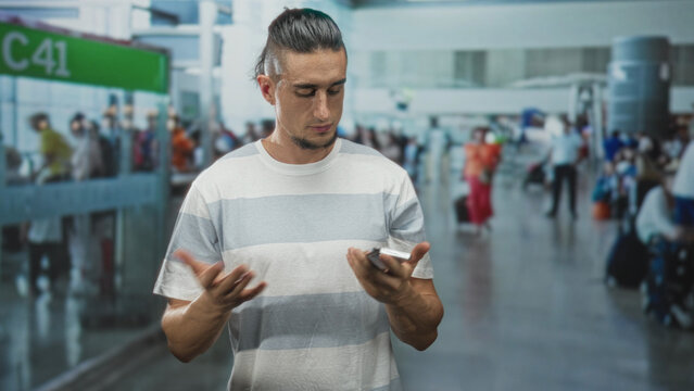 Man in white and gray striped t shirt taps smartphone screen and sometimes holds phone to ear in airport; waiting focus.