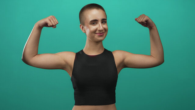 Woman flexing biceps with clenched fists against a green studio wall, short hair visible and toned arms on display; confidence empowerment.