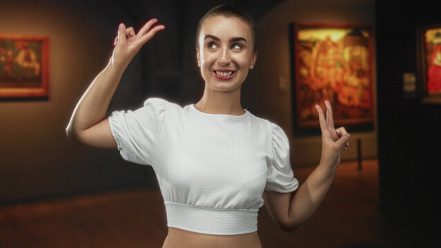Woman with short hair flashing peace signs with both hands and visible fingers while smiling in a museum gallery with framed paintings and spotlights; playful joy.